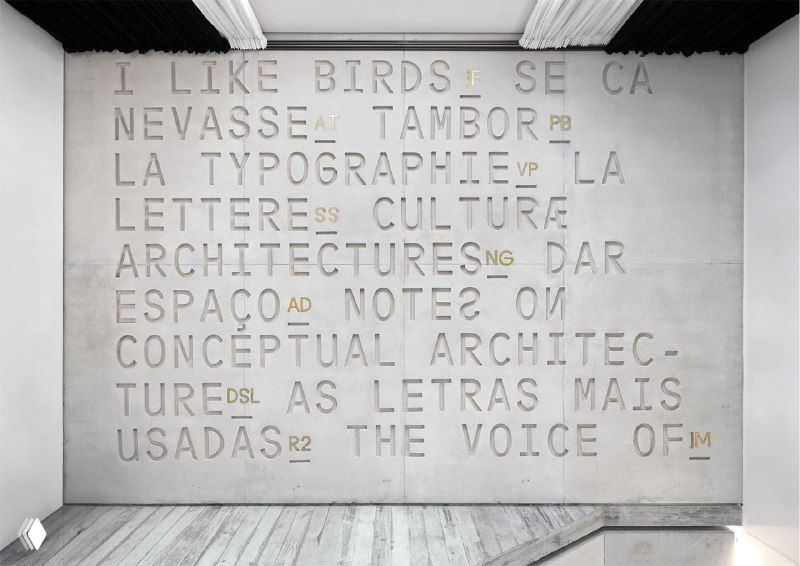 Широкая стена с крупной рельефной типографикой: ровные строки больших букв на бетонной поверхности в минималистском интерьере.