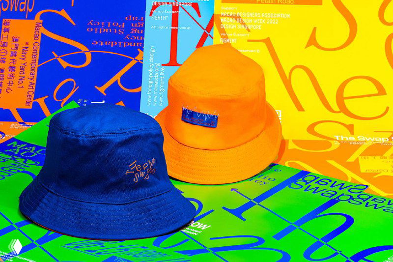 Два панамы (синяя и оранжевая) на фоне графических плакатов The Swap Show с контрастной типографикой и кислотными цветами.
