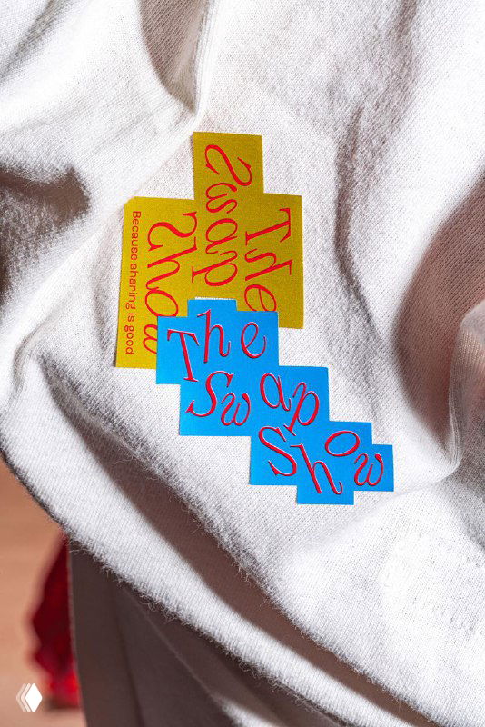 Белая футболка с нашивкой в виде цветного пазла и надписью The Swap Show, крупный план текстуры ткани и стилизованного принта.