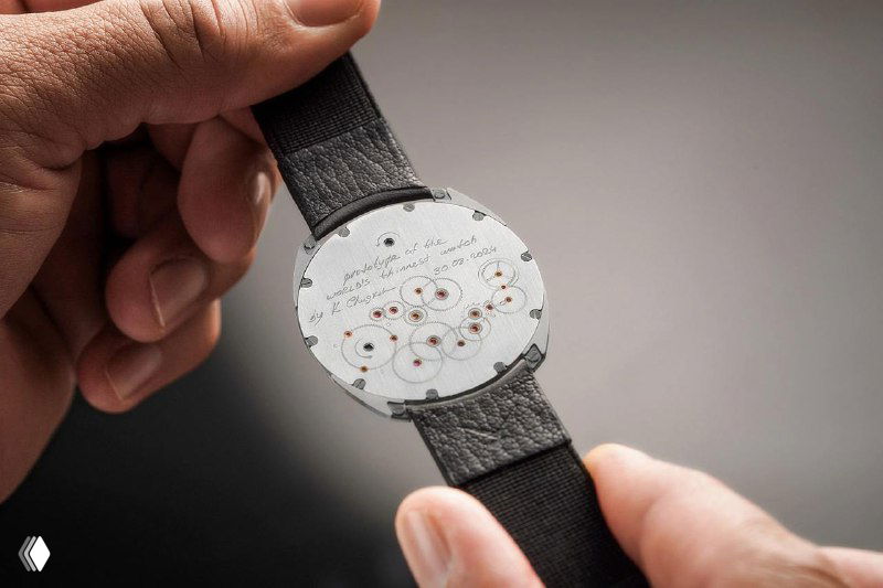 Крупный план: мастер держит платину механизма часов Thinking, видно множество мелких деталей и отверстий для элементов механизма, аккуратная сборка.