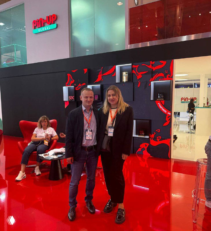 Two attendees posing in front of a bright red exhibition booth at SBC Summit Lisbon, both wearing badges and smiling.