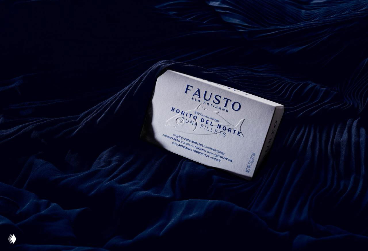 Упаковка Fausto в перспективе на тёмном фоне: коробка с тиснением, контраст между белым фактурным картоном и насыщенно-синим окружением ткани.