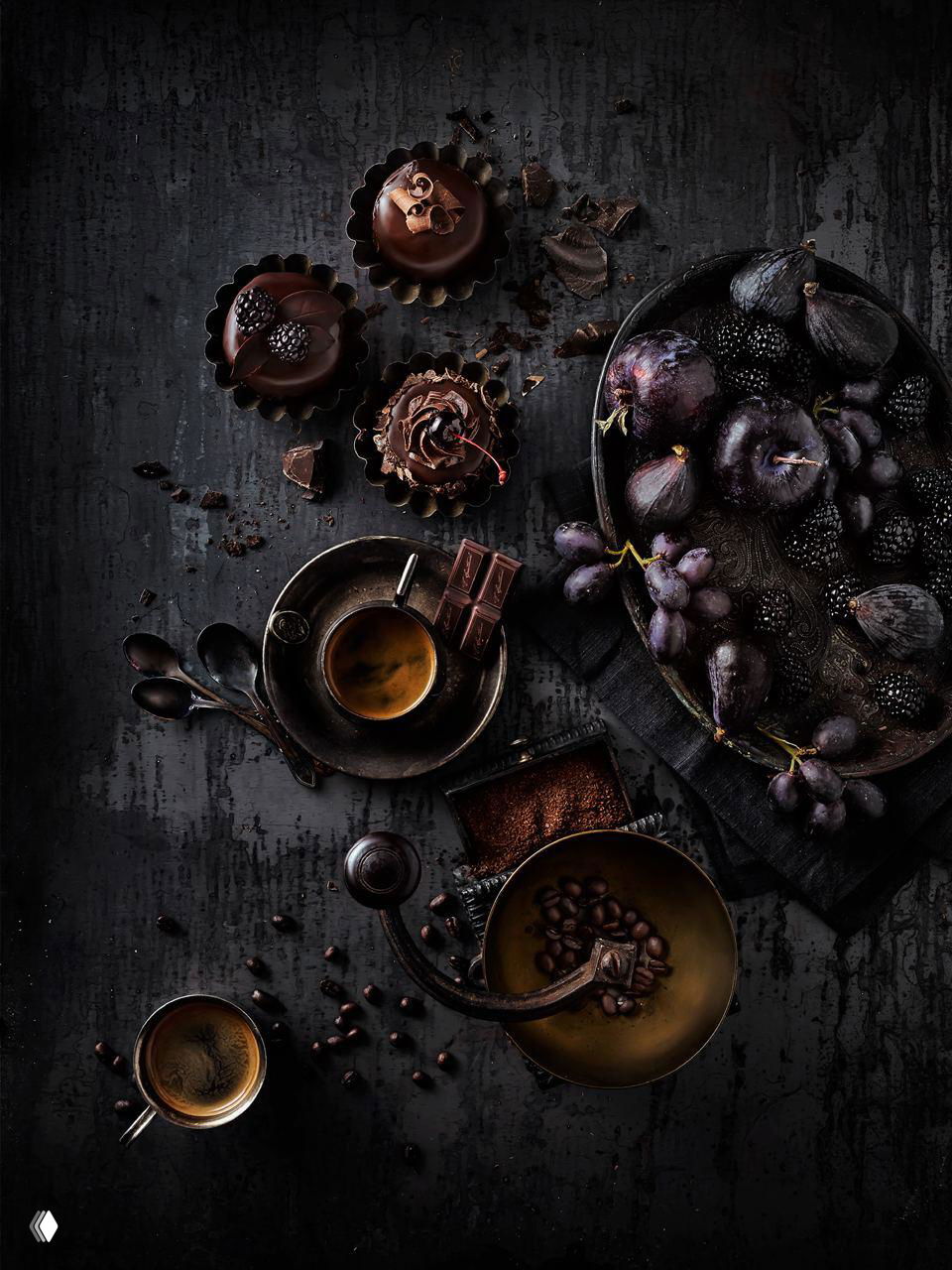 Тёмная стилизованная фуд-фотография: чашки кофе, шоколадные тарталетки, инжир и зерна кофе на грубой тёмной поверхности в художественной композиции.
