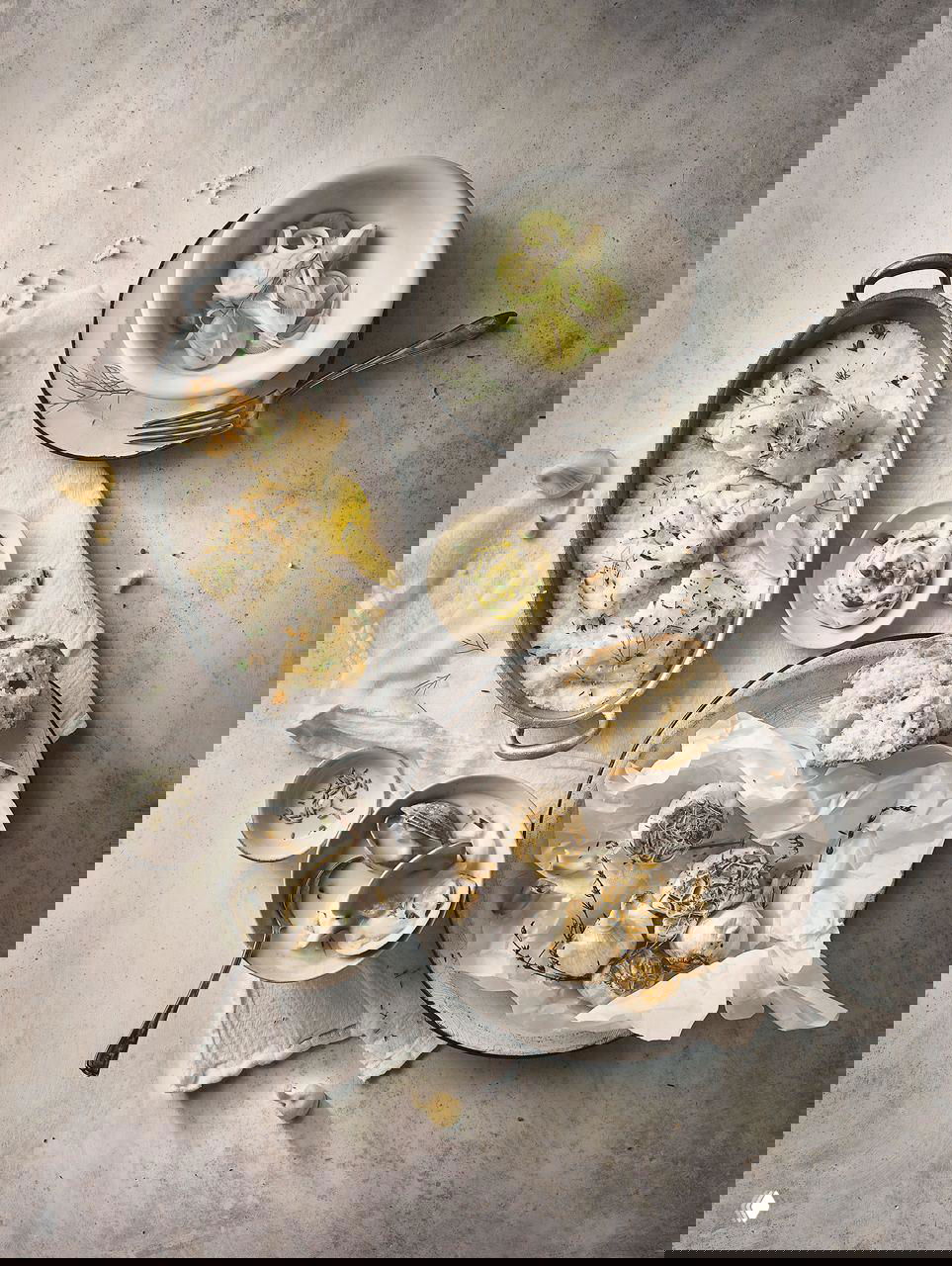 Светлая фуд-фотография: тарелка с запечённой рыбой, чесноком и лимоном, рядом хлеб и кремовый соус на светлом фоне в аккуратной натюрморт-композиции.