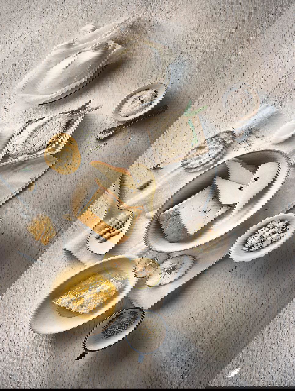 Минималистичная фуд-съёмка в белых тонах: сыр, груша, мёд и крекеры на светлом деревянном фоне, аккуратно разложенные предметы и текстуры.