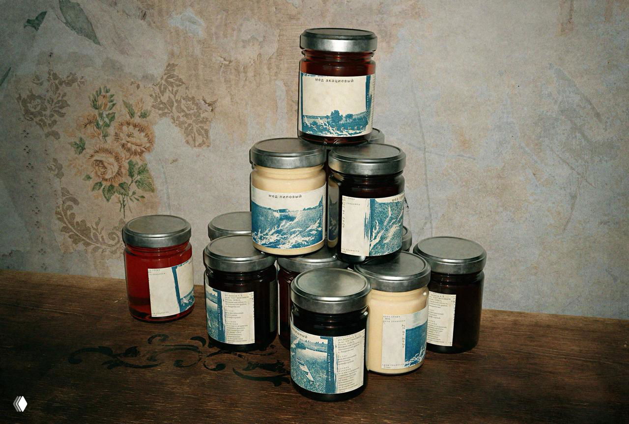 Пирамида небольших баночек с мёдом и настойками на деревянной поверхности, этикетки с растительными отпечатками и текстом.