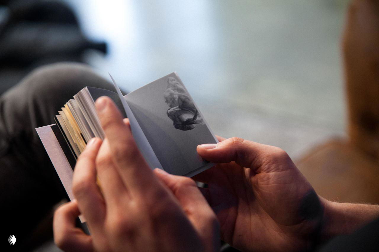 Руки держат мини‑книгу с синими и белыми графическими элементами; кадр подчёркивает фактуру бумаги и плотность блока страниц.