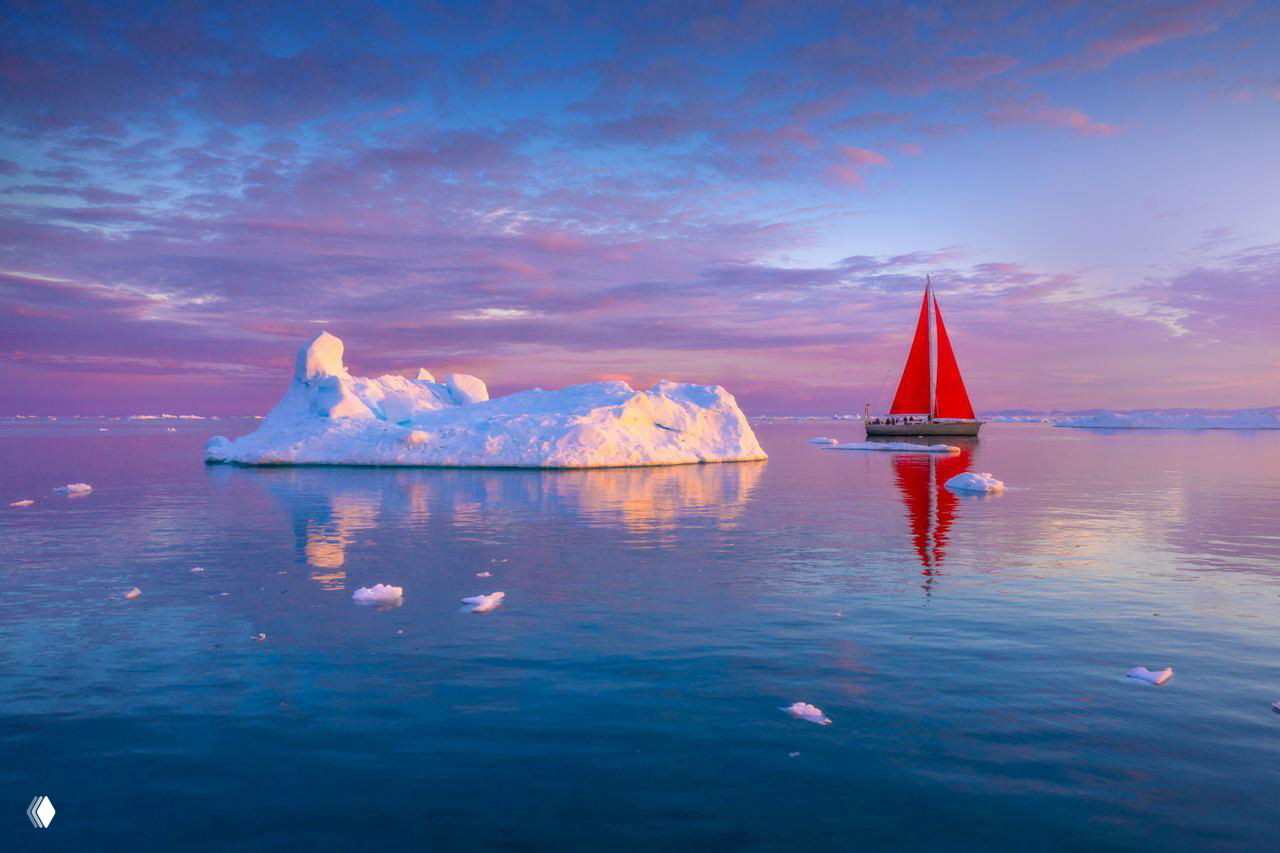 Парусник с ярко‑красными парусами на спокойной зеркальной воде рядом с крупным айсбергом в пастельном свете Диско‑Бэй, Гренландия.