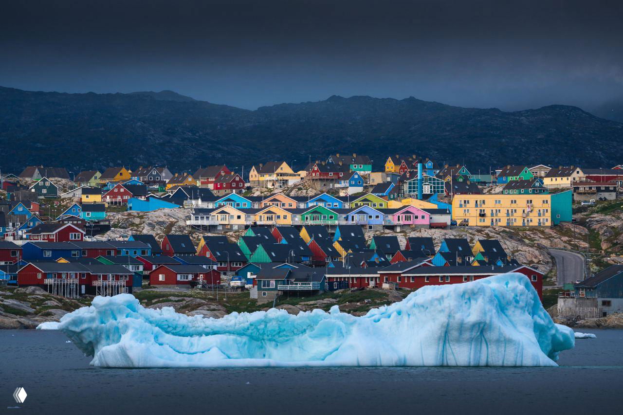 Прибрежный поселок с яркими разноцветными домами на скалистом берегу и крупный айсберг на воде у города в холодном освещении.