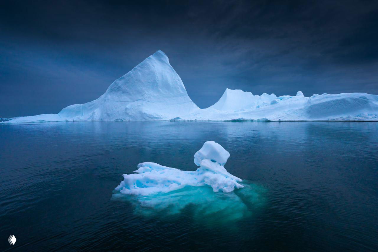 Большой айсберг с выразительным силуэтом и сложной формой на фоне тёмного неба и гладкой морской поверхности у берегов Гренландии.