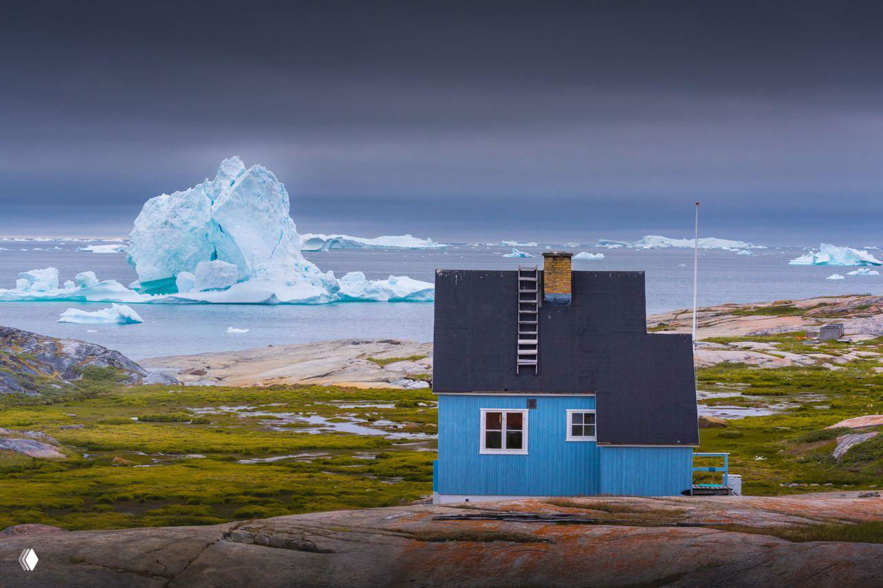 Дом у моря голубого цвета на мхистых скалах с айсбергами на горизонте, сценка прибрежной жизни и одиночества северной деревни.