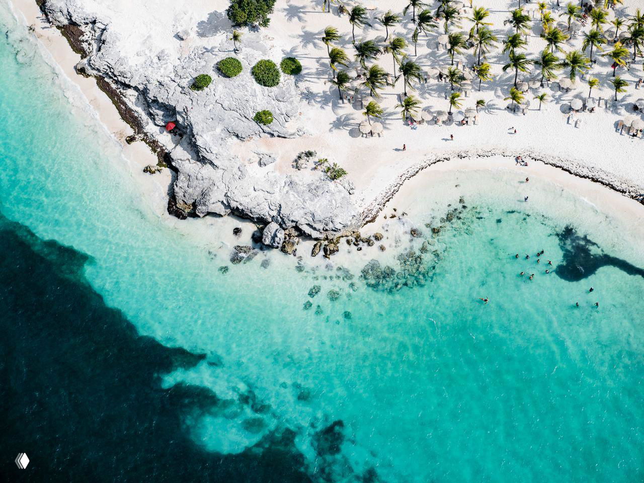 Аэрофото карибского побережья: контраст тёмных вод и бирюзовых мелей, белый песок, скальные выходы и редкие люди на пляже в кадре сверху.