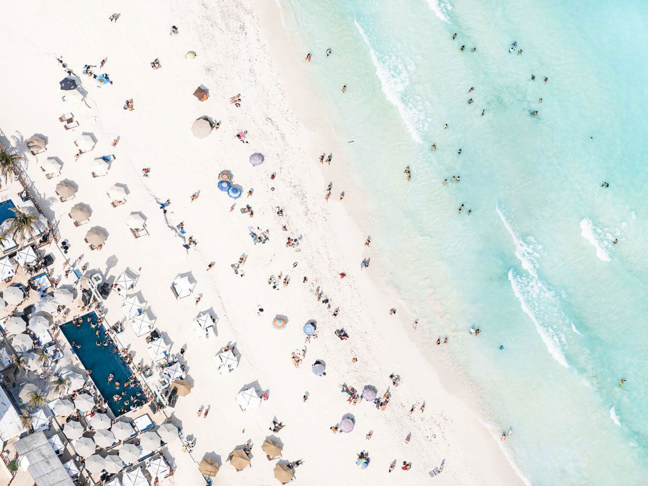 Снимок пляжной зоны сверху с зонтиками, лежаками и группами людей; белый песок переходит в прозрачную бирюзовую воду.