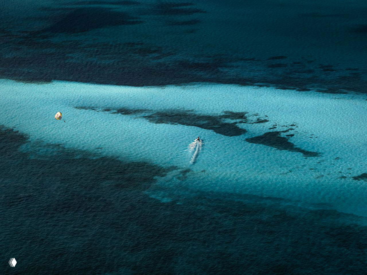 Крупный аэрокадр с одинокой моторной лодкой и тёмными подводными участками, подчёркнутыми на фоне ярко-голубой поверхности.