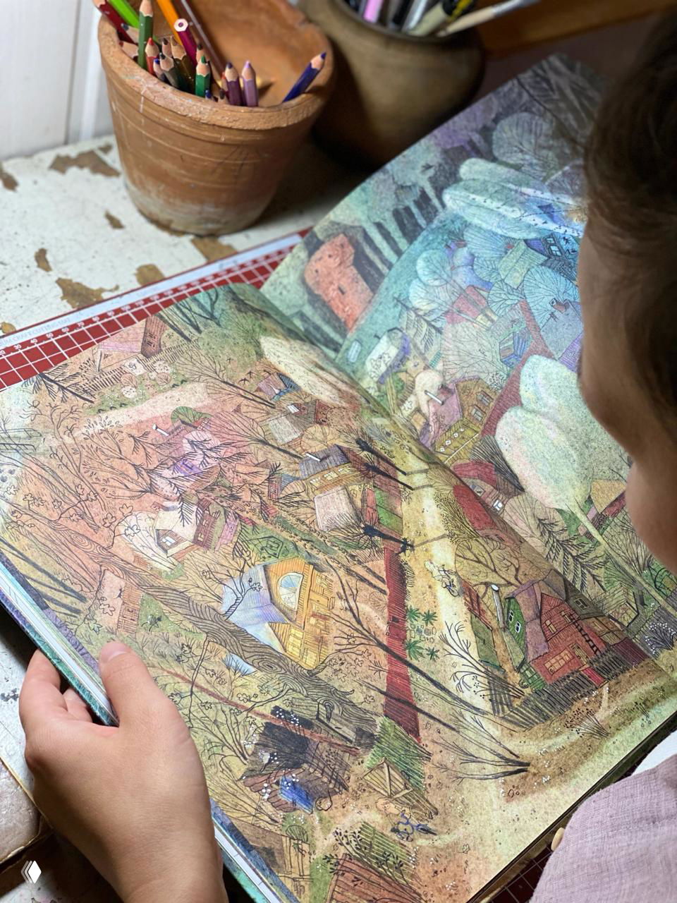 Фотография разворота книги в руках ребёнка: крупный план детализированной иллюстрации деревни, заметна фактура бумаги и цветные карандаши на столе.