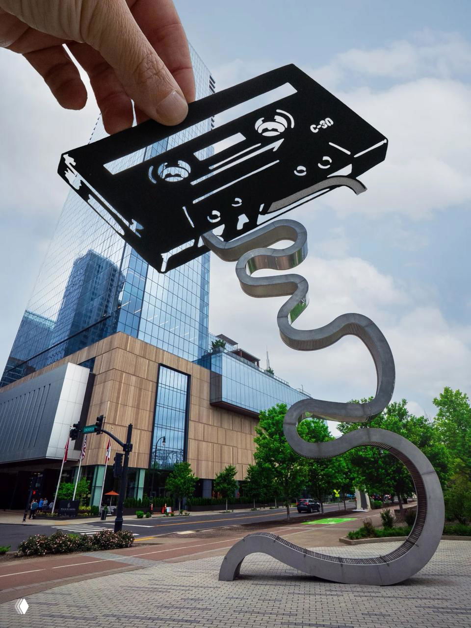 Рука помещает чёрно‑белую вырезанную аудиокассету над современной городской скульптурой, создавая визуальную ироничную связь.