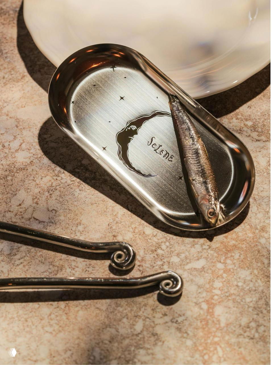 Небольшой металлический поднос с тиснёным полумесяцем и надписью Selene, рядом селёдка; отражения и приглушённый свет подчёркивают материал и текстуру.