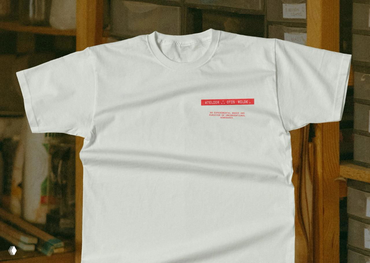 Белая футболка с небольшим красным принтом A.Otis на фоне мастерской: мерч в фирменной стилистике студии, демонстрация брендинга в носителях.