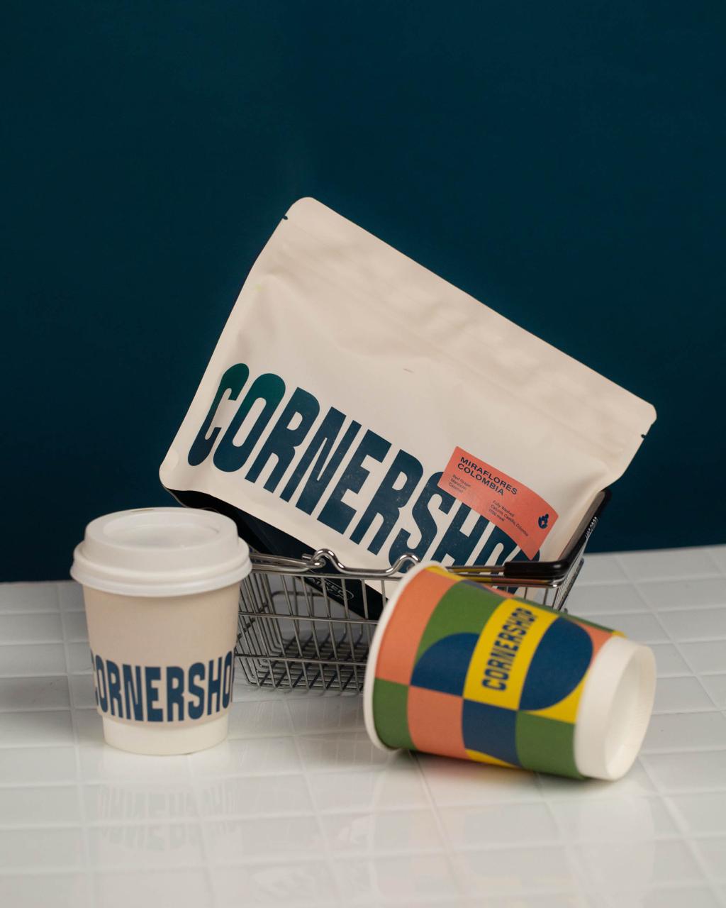 Упаковка кофе с логотипом Cornershop и два брендированных стаканчика в корзинке на плиточной поверхности на тёмно-зелёном фоне
