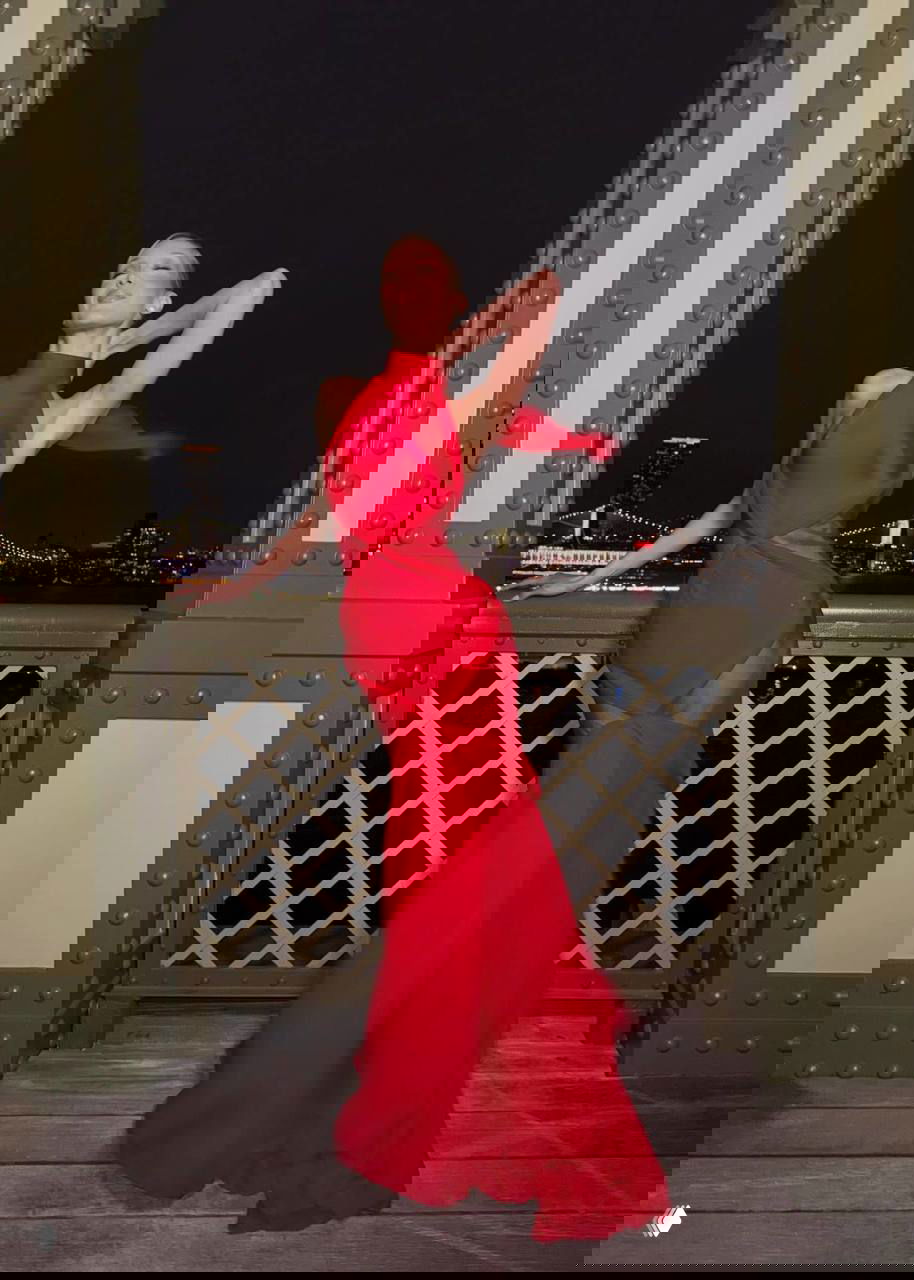 Белла Хадид в красном платье Schiaparelli позирует на ночной набережной: прозрачные вставки, струящийся шлейф и огни города позади.