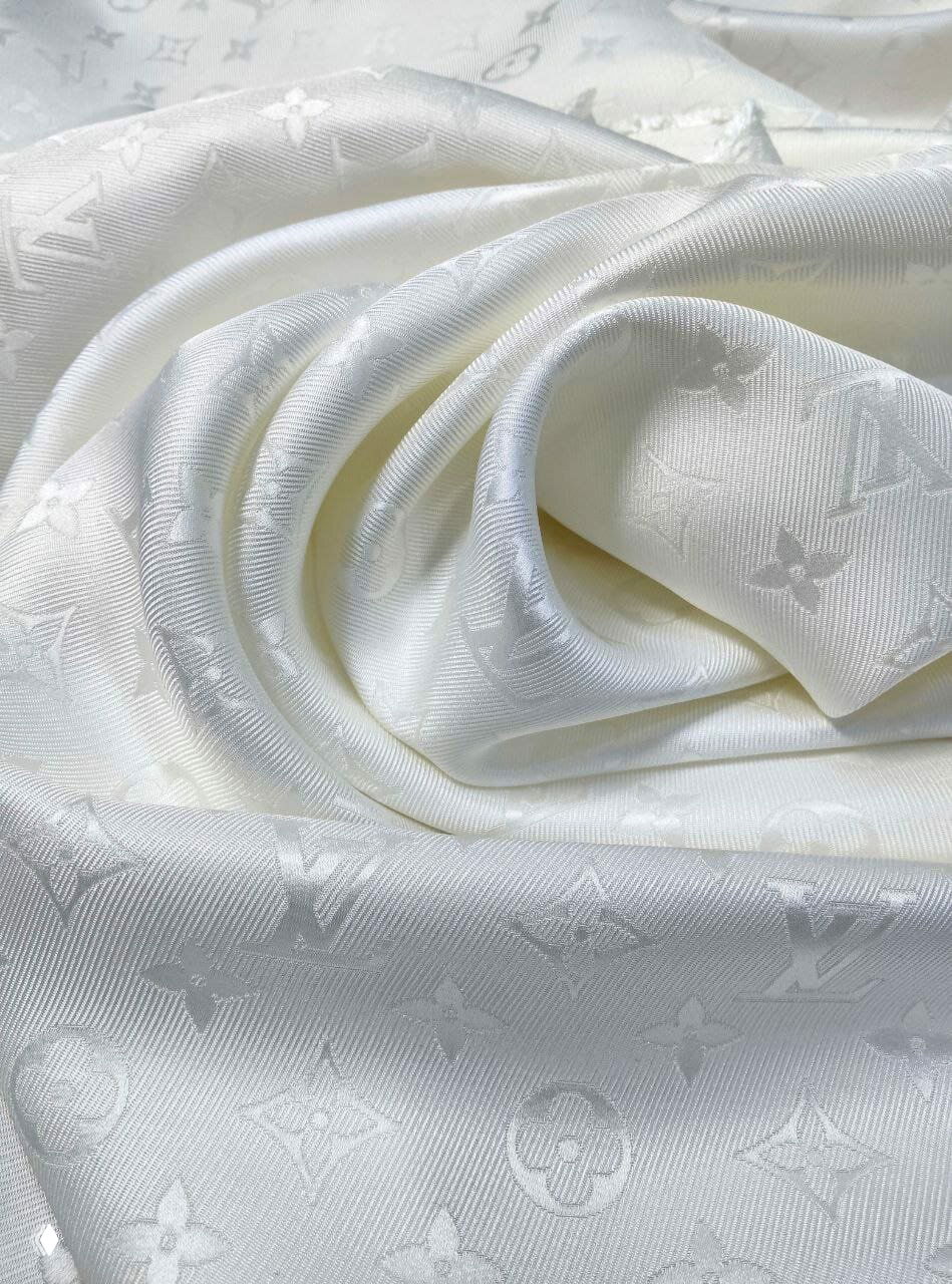 Крупный план белой жаккардовой ткани с тиснённым узором в виде повторяющихся флористических и монограммных мотивов, гладкая текстура.