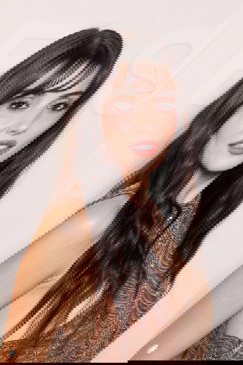 Крупный кадр лица Ханде Эрчел с лёгким румянцем и чёлкой, видны детали макияжа, ресниц и текстура украшенного топа на фоне логотипа Elie Saab.