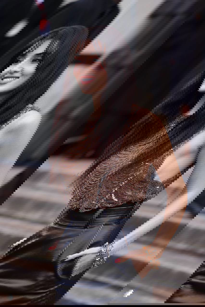 Ханде Эрчел в движении возле здания показа: коричневый топ, кожаная юбка, боковой профиль и счастливая улыбка; видны детали образа и массивный браслет на руке.