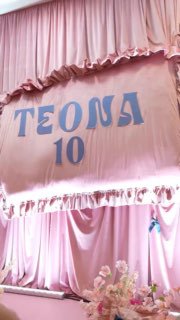 Розовый занавес сцены с крупной надписью TEONA 10 и цветочной композицией у подножия, видна праздничная декорация зала
