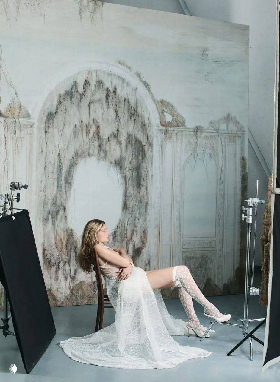 Модель сидит на стуле в прозрачном кружевном платье и высоких кружевных чулках на фоне художественной декорации студии.