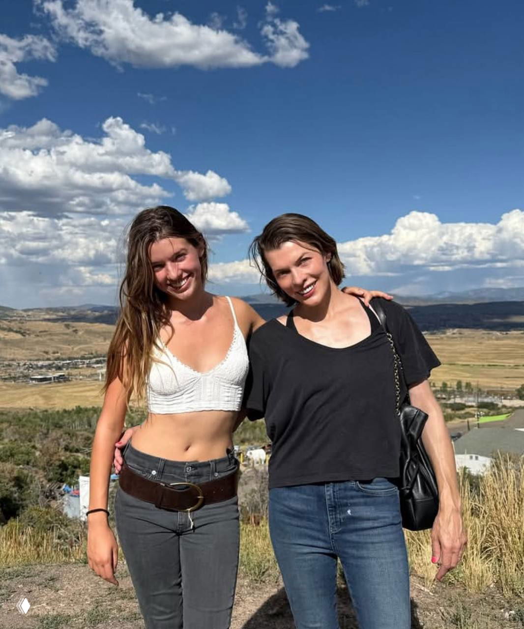 Женщина и молодая девушка стоят на фоне широкого ландшафта и неба с облаками; летний уличный кадр, непринуждённая поза и улыбки.