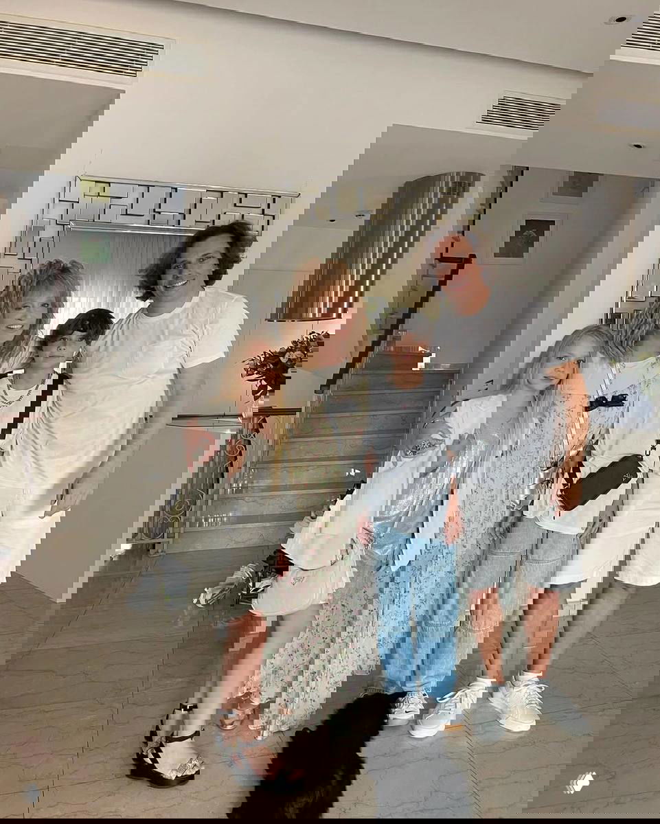 Фотография в интерьере виллы: пожилая женщина и трое человек позируют в светлом холле на фоне лестницы и зеркала.