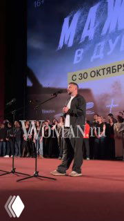 Человек на сцене с микрофоном перед большим экраном, на экране видна рекламная графика с текстом и датой релиза фильма.