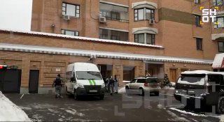 Вход в жилой дом зимой: у порога стоят служебные фургоны и несколько людей, снег на земле, уличная обстановка.