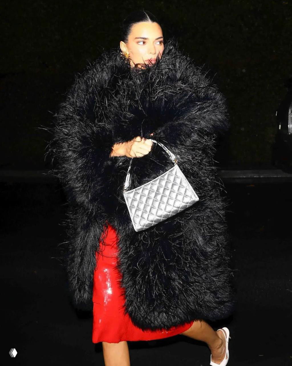 Фото папарацци: женщина укутана в чёрную объёмную шубу, видна часть красного платья и серебристая сумка — контрастные фактуры и аксессуары.