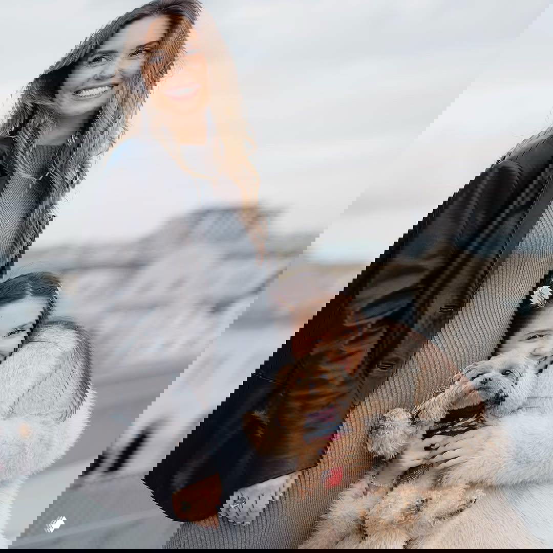 Агата Муценице улыбается рядом со старшей дочерью Мией, которая крепко обнимает маленькую собачку на фоне городской панорамы и осеннего неба.