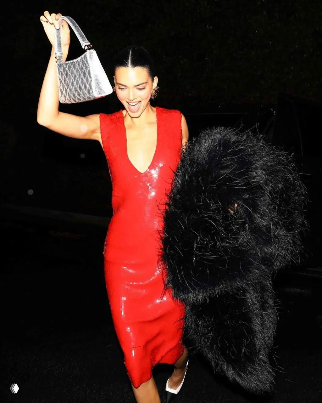 Динамичный снимок: женщина в красном платье поднимает руку с сумкой и улыбается, папарацци-стиль съёмки на тёмной улице ночью.