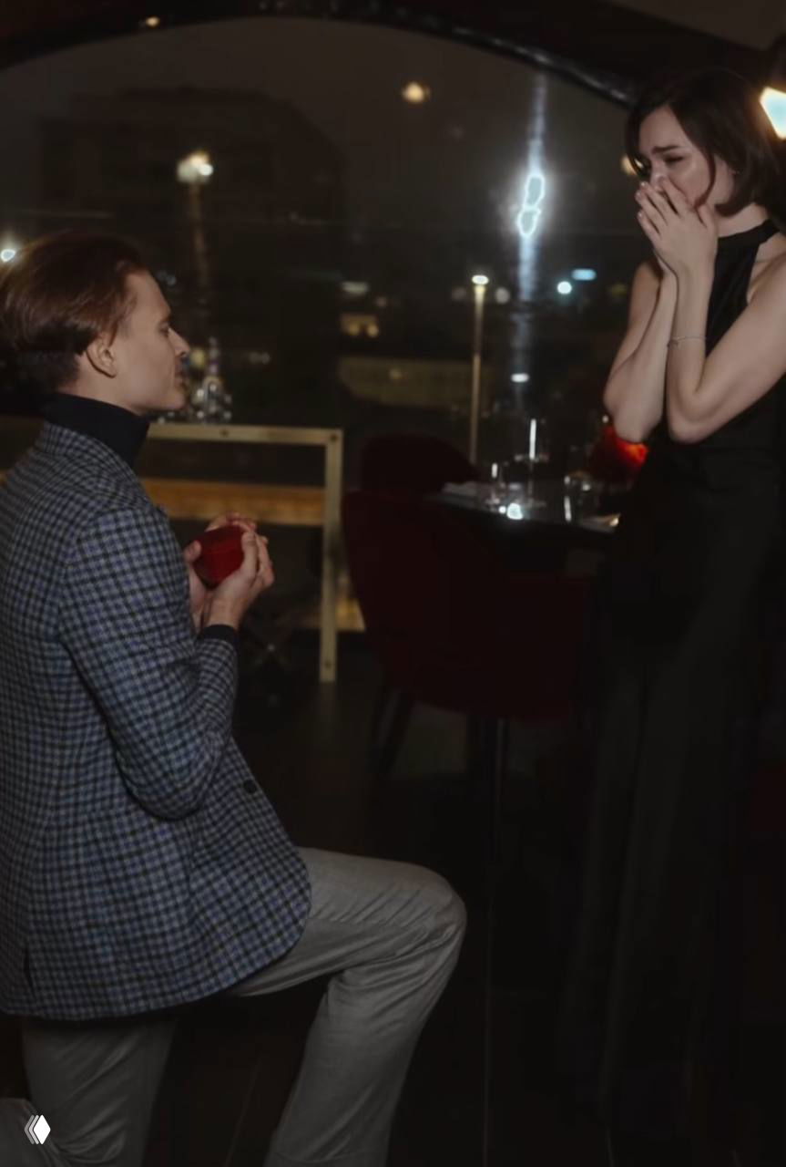 Момент предложения: мужчина на колене держит раскрытую шкатулку с кольцом; перед ним Евгения Медведева закрывает рот руками в удивлении.