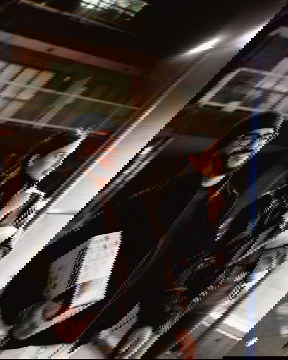 Тимати в тёмной шапке и женщина в кожаной куртке идут по ночной улице; он несёт коробку, вокруг урбанистический фон и искусственное освещение.