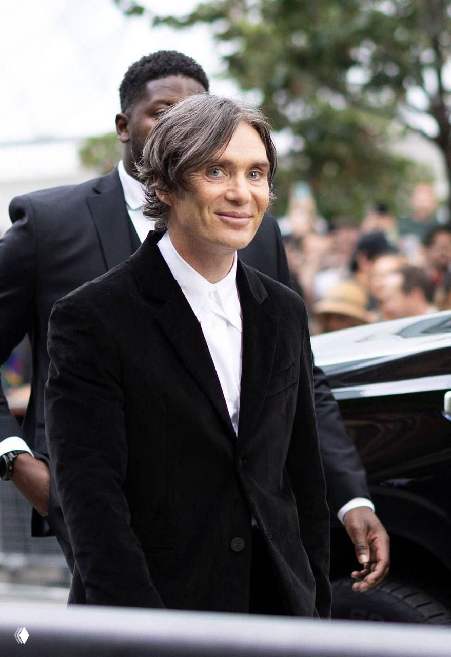 Цветная фотография мужчины в чёрном пиджаке и белой рубашке на улице у автомобиля, люди и охрана на заднем плане