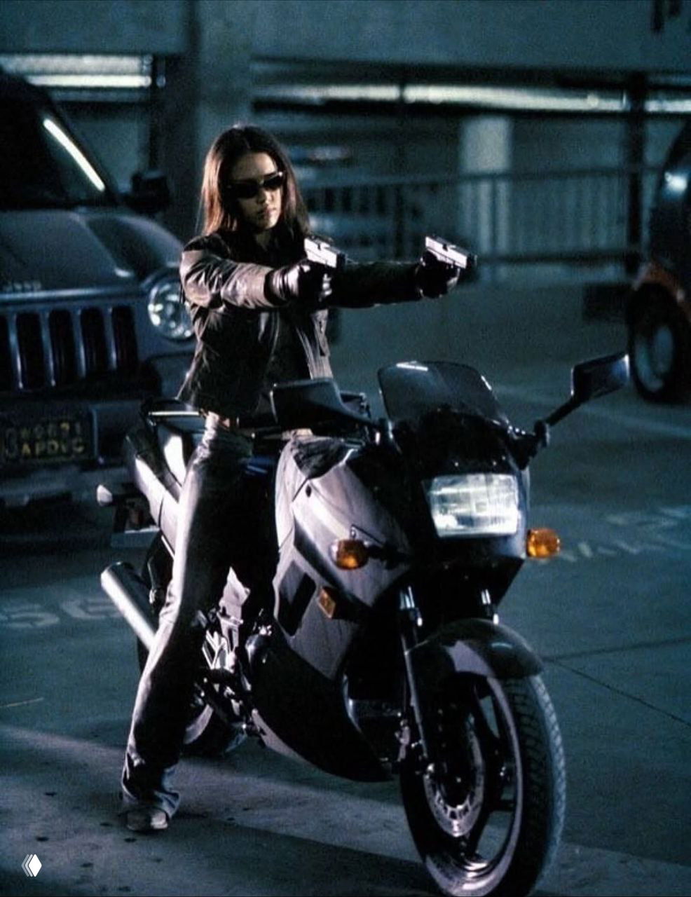 Кадр из сериала: Джессика Альба стоит возле мотоцикла и держит два пистолета, динамичная сцена в городской парковке ночного времени.