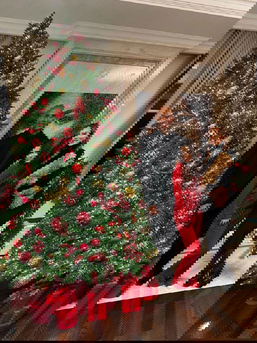 Высокая рождественская ёлка с золотыми и красными шарами, вокруг подарочные коробки и семья на фоне — праздничная интерьерная композиция.