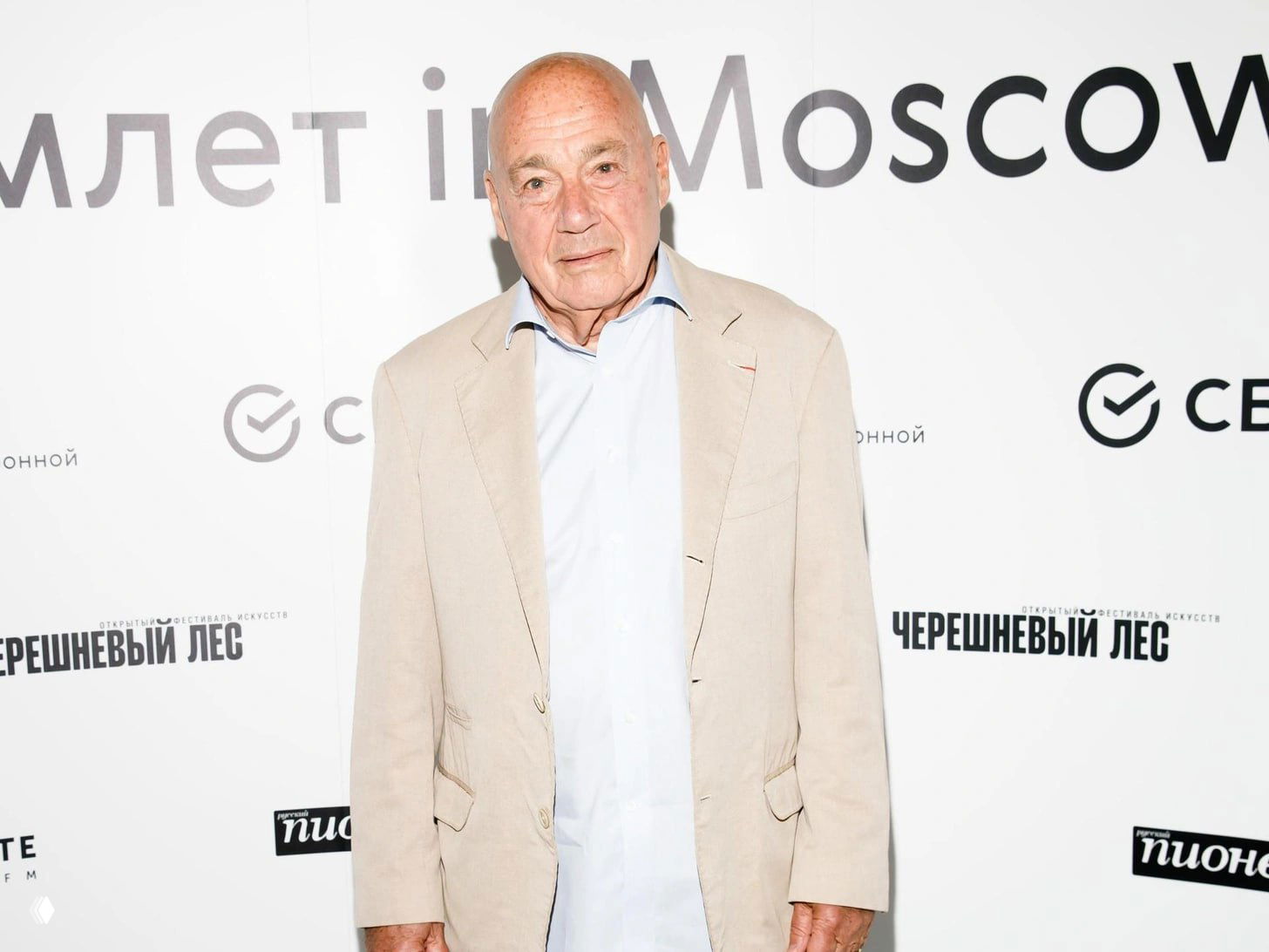 Портрет пожилого мужчины в светлом пиджаке на пресс‑волле, студийная съёмка на светлом фоне с логотипами и минимальными тенями.