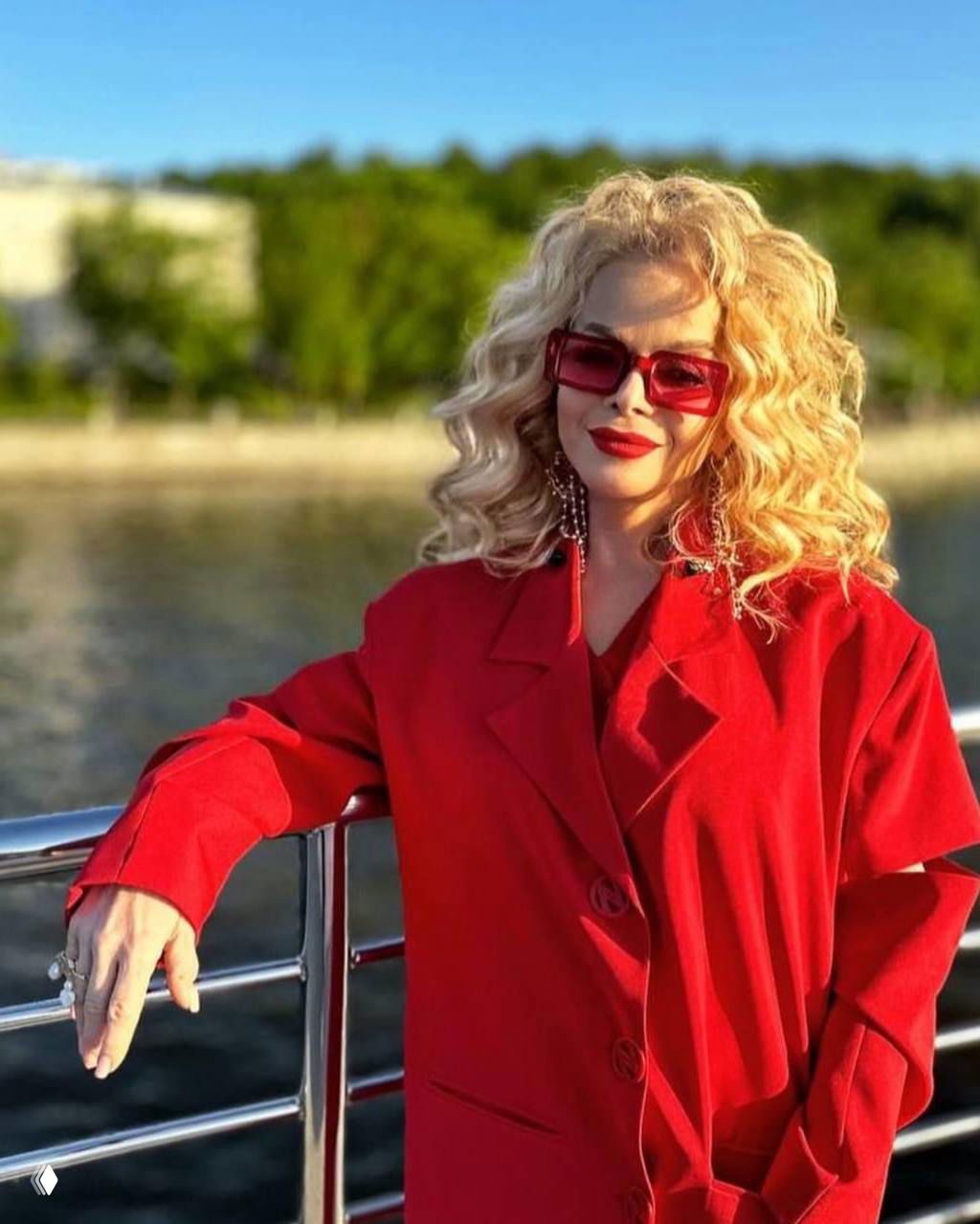 Певица Лариса Долина в ярко‑красном пальто и крупных розовых очках позирует у перил на фоне реки и зелёной набережной.
