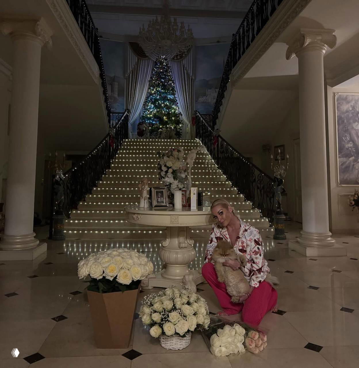 Широкий холл с подсвеченной лестницей, центральный стол с букетами белых роз и большая ёлка в глубине — роскошный праздничный интерьер.
