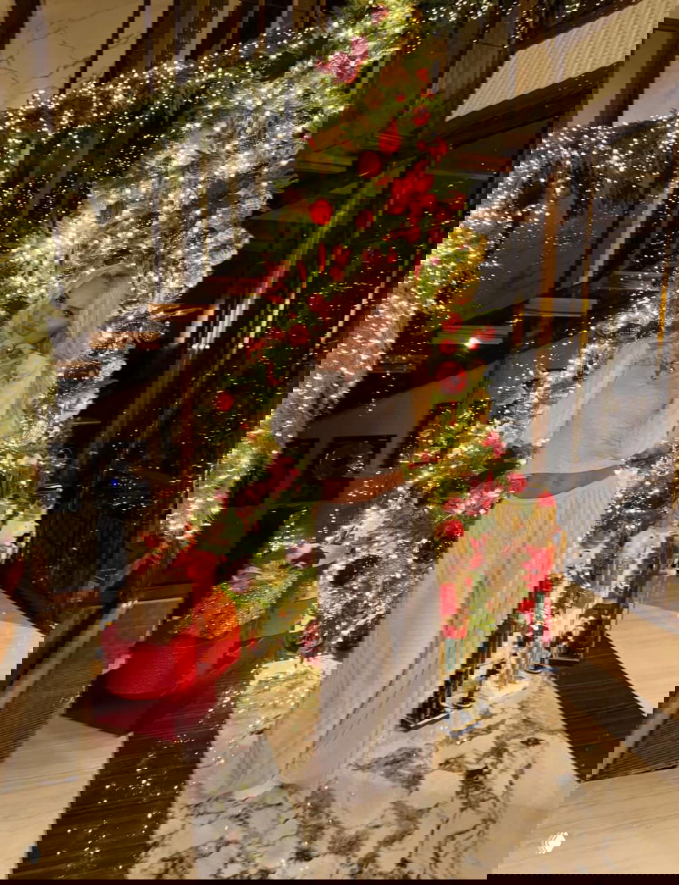 Молодая женщина позирует у украшенной светящейся ёлки в уютной зоне дома, рядом — декоративные музыкальные солдаты и подарки на полу.