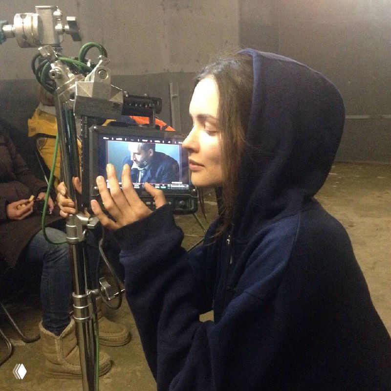 Паулина Андреева в тёплой толстовке и капюшоне смотрит на монитор съёмочного кадра — закулисный момент проверки сцены и работы команды.