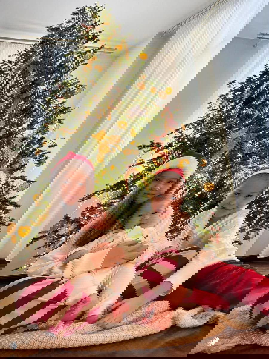 Три ребёнка в праздничной одежде сидят на полу перед украшенной ёлкой: красно-белые пижамы и милые рождественские детали в окружении.