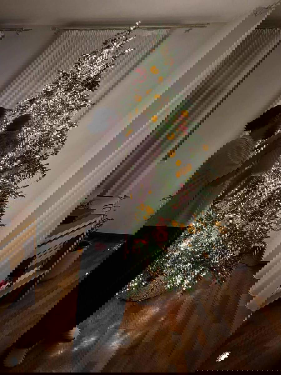 Отец держит ребёнка на руках перед украшенной гирляндами ёлкой в темной комнате — мягкая подсветка создаёт атмосферу уюта и тепла.