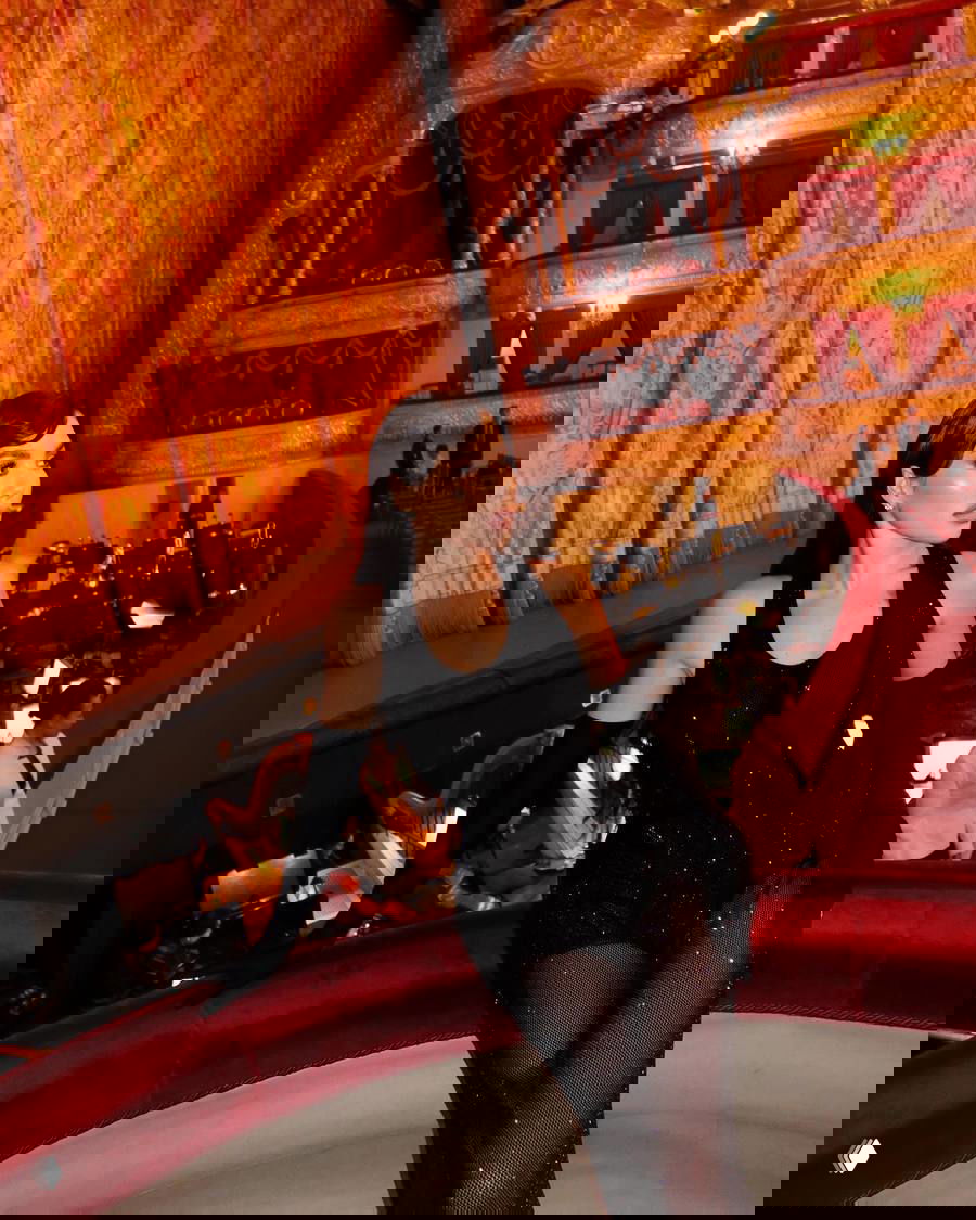 Женщина в чёрном блестящем платье и длинных перчатках позирует в ложe Большого театра на фоне золотого занавеса и оркестровой ямы.