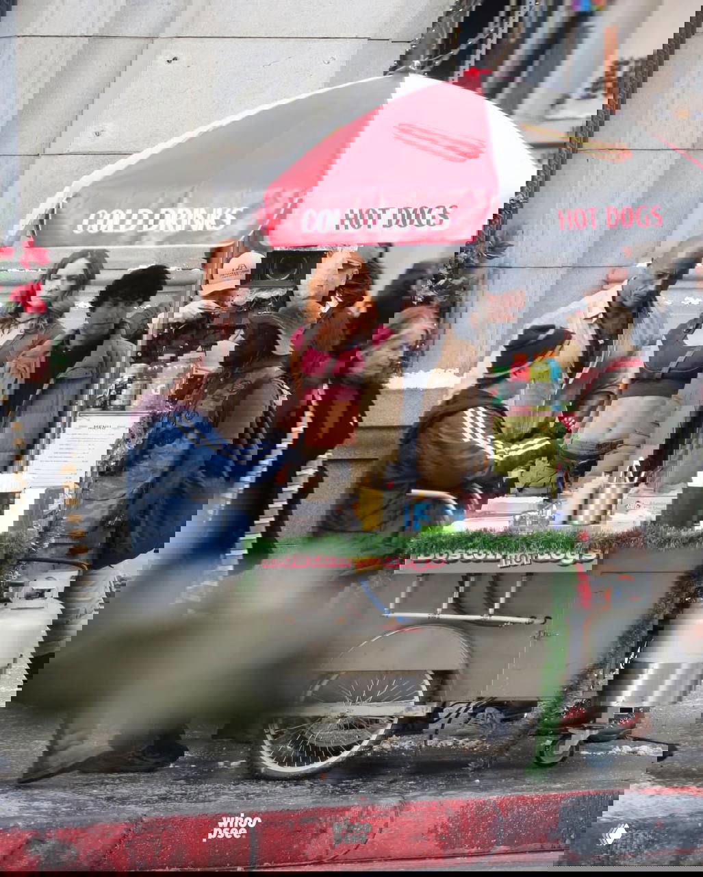 Съёмочная сцена на улице: несколько актёров у тележки с хот‑догами, украшенной гирляндой и искусственным снегом вокруг.